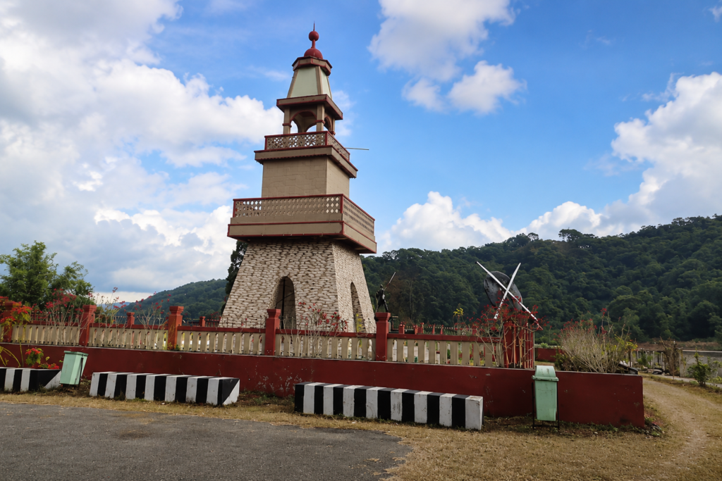 A tall, beige tower with red trim stands in a fenced area by a road, set against green hills and a blue sky with clouds. Shrubs and decorative plants surround the fence.