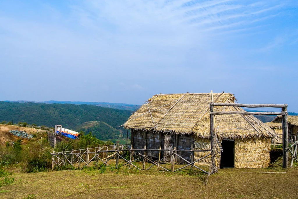 A thatched-roof hut with wooden walls stands on dry ground, surrounded by a simple bamboo fence, with green hills and a blue sky in the background.