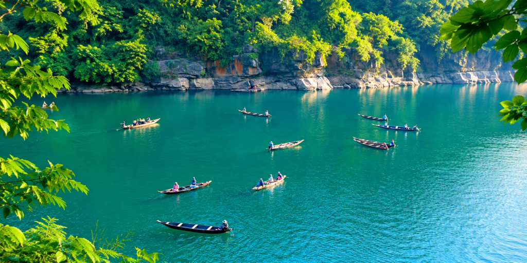 Boats with people glide on clear turquoise water, surrounded by lush green trees and rocky cliffs, under bright daylight.
