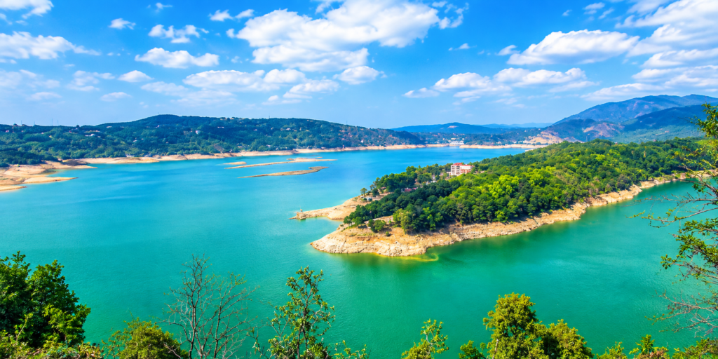 Turquoise lake surrounded by lush green hills and mountains under a bright blue sky with scattered clouds. Small peninsulas covered in trees extend into the water.