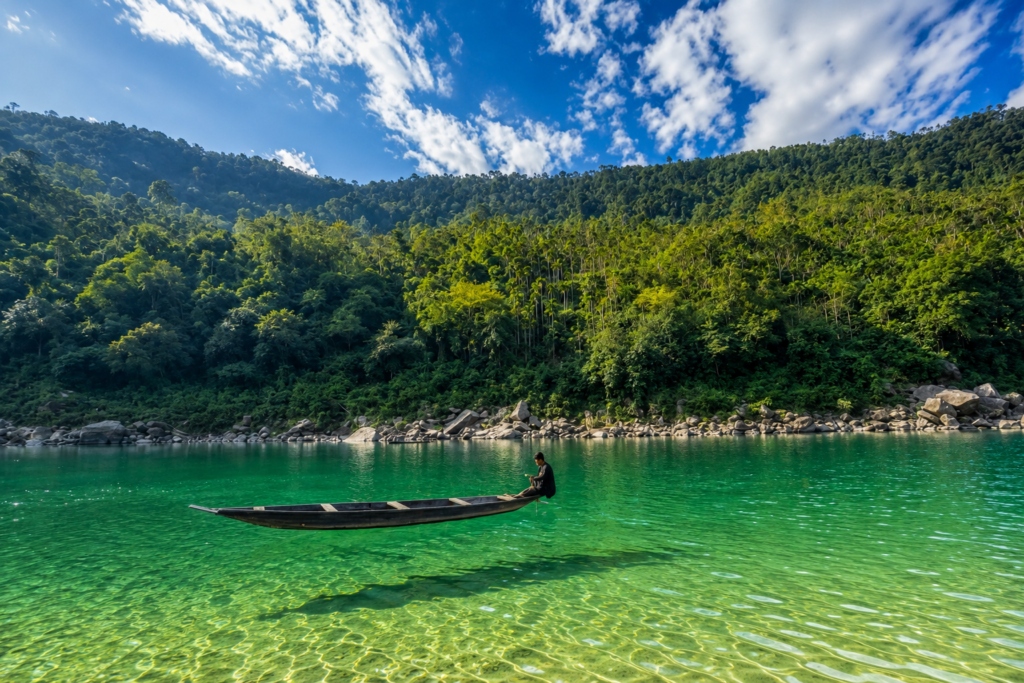 A person sits in a wooden canoe on clear turquoise water, surrounded by lush green forested hills under a blue sky with scattered clouds.