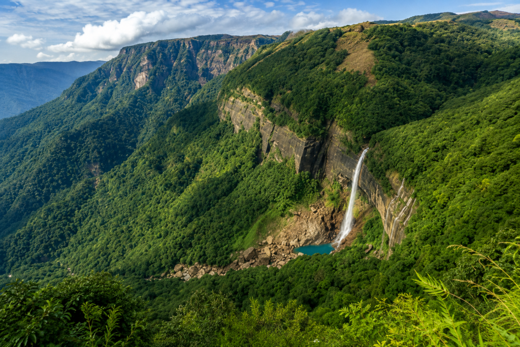 A tall waterfall cascades down a lush green mountainside into a blue pool, surrounded by dense forest and cliffs under a partly cloudy sky.