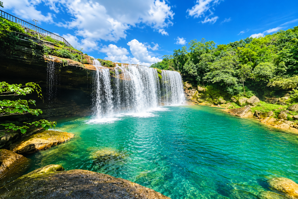 A beautiful waterfall cascades into a clear turquoise pool surrounded by lush green trees, rocks, and a bright blue sky with scattered white clouds.