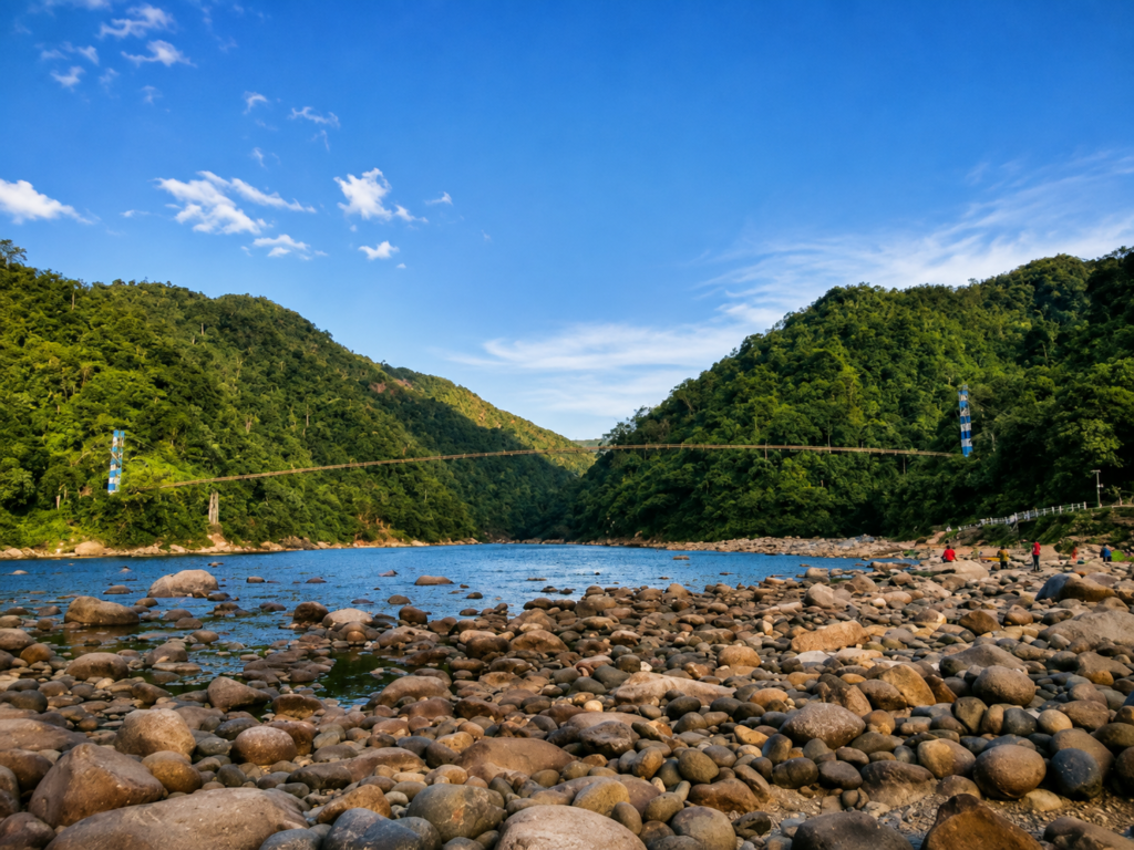 Rocky riverbank with clear water, surrounded by lush green hills. A long suspension bridge spans the river, under a bright blue sky with scattered clouds. People are walking near the waters edge.
